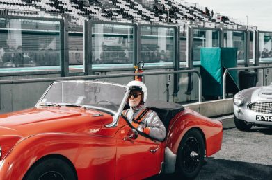 Un homme en combinaison de course grise et avec un casque de pilote blanc est assis dans une voiture de course classique rouge, prêt à prendre la piste. Il porte des gants de course et une ceinture de sécurité tout en se concentrant sur la piste devant lui. Derrière lui, des spectateurs sont visibles à travers la fenêtre réfléchissante d'une loge portant le logo d'AUTOSUR.