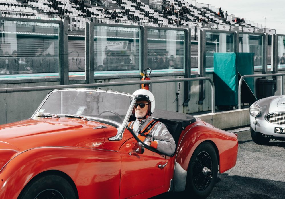 Un homme en combinaison de course grise et avec un casque de pilote blanc est assis dans une voiture de course classique rouge, prêt à prendre la piste. Il porte des gants de course et une ceinture de sécurité tout en se concentrant sur la piste devant lui. Derrière lui, des spectateurs sont visibles à travers la fenêtre réfléchissante d'une loge portant le logo d'AUTOSUR.